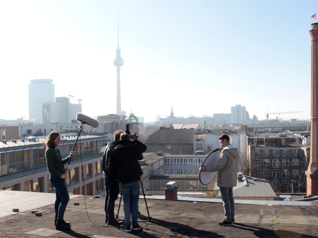 : Im Vordergrund dreht eine 5-köpfige Gruppe auf einem Dach einen Film. Links hält eine Person ein großes Mikro, eine andere Person rechts einen Reflektor, in der Mitte stehen zwei Personen hinter und eine vor der Kamera. Im Hintergrund befindet sich die Stadtsilhouette Berlins einschließlich des Fernsehturms an einem sonnigen, wolkenlosen Tag. 